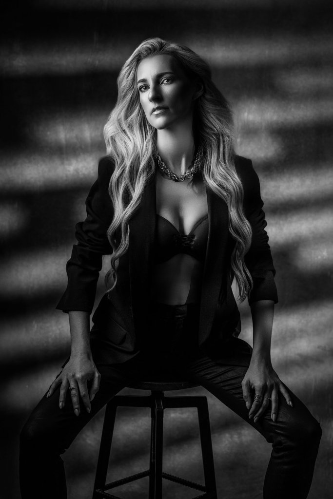 Czarno-biała sesja sensualna – blondynka z długimi falami włosów w marynarce na biustonoszu, na stołku