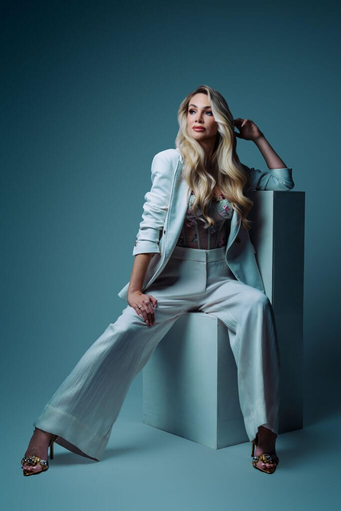Elegancka sesja kobieca – blondynka w jasnym garniturze i koronkowym topie na tle w kolorze błękitu